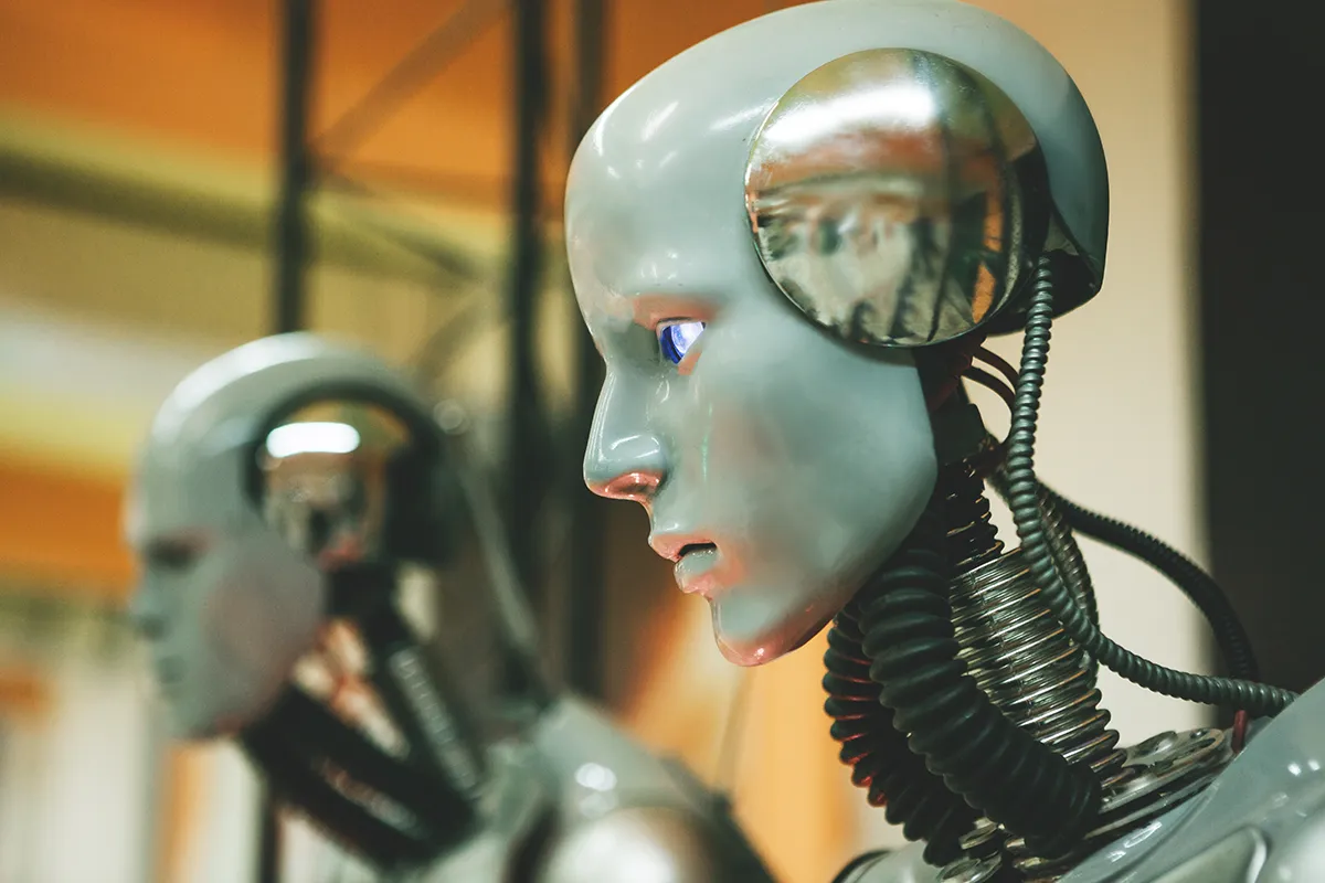 image depicting a robot being built as part of a group of Technology Investments conducted in america, robot has a white head and exposed wire to the torso, there are two robots one in focus and one not